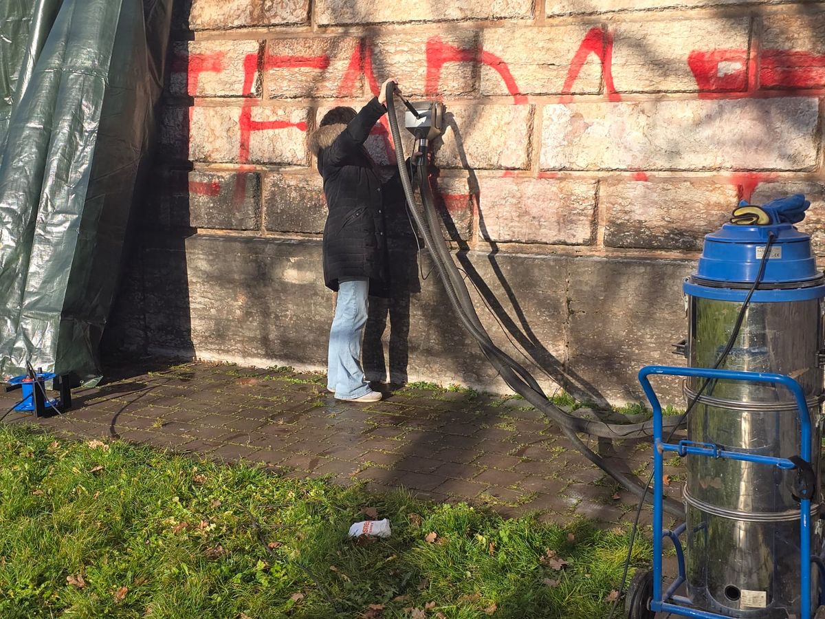 Reinigung eines rot besprühten Sandsteinquaders am Denkmal, Abdeckplane schützt die Umgebung